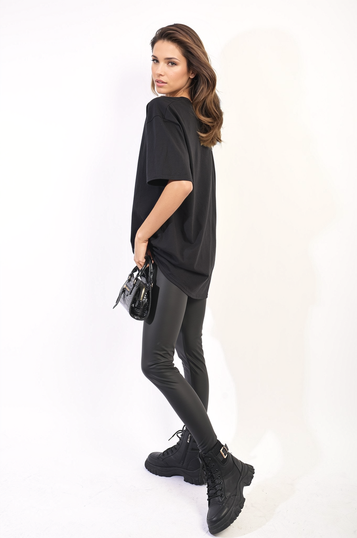 Woman wearing the Amanda Oversized Love Slogan Print Top with black leggings and boots, holding a purse. Perfect for casual, stylish looks from Holiday-clothes.co.uk.