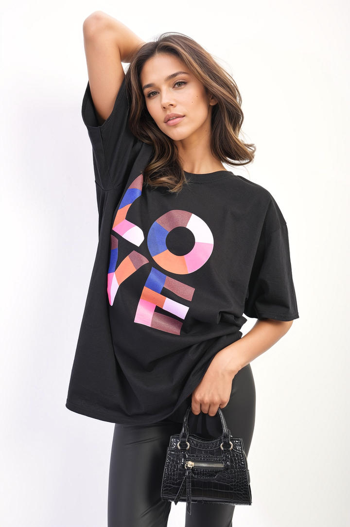 Woman wearing Oversized Love Slogan Print Top, posing with black purse. The top features a bold graphic design, offering a stylish and relaxed look.