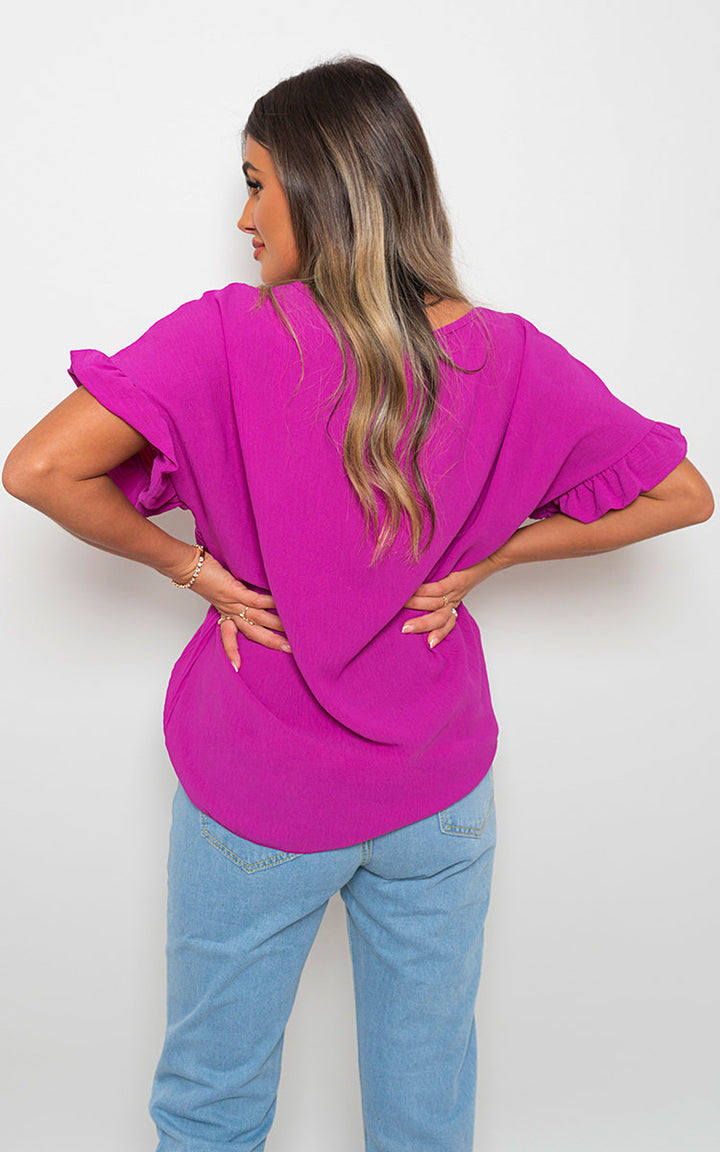 Woman wearing Bella Frill Sleeve Top With Chain, showcasing its stylish frill sleeves and chain detail, paired with blue jeans for a chic look.