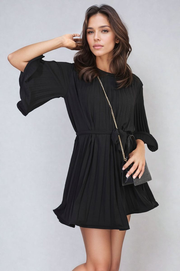 Woman in black pleated tie waist mini dress with long brown hair, holding a purse, exemplifying chic elegance. Perfect for versatile styling.