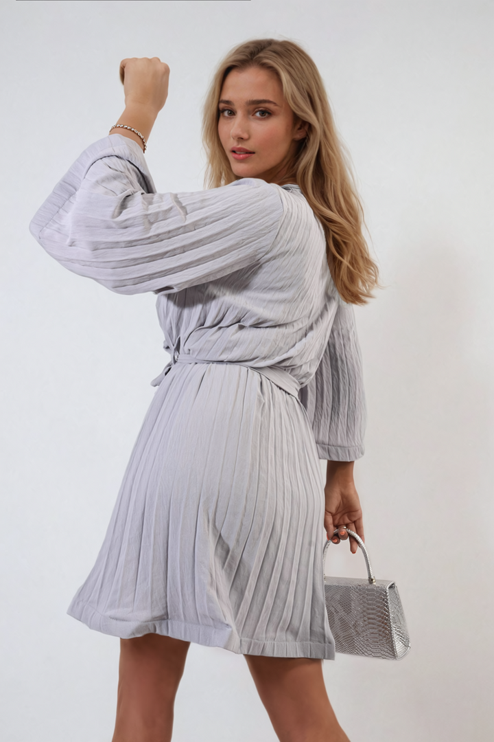 Woman modeling Carmen Pleated Tie Waist Mini Dress, holding a silver bag, showcasing elegant pleating and a flattering fit for stylish occasions.