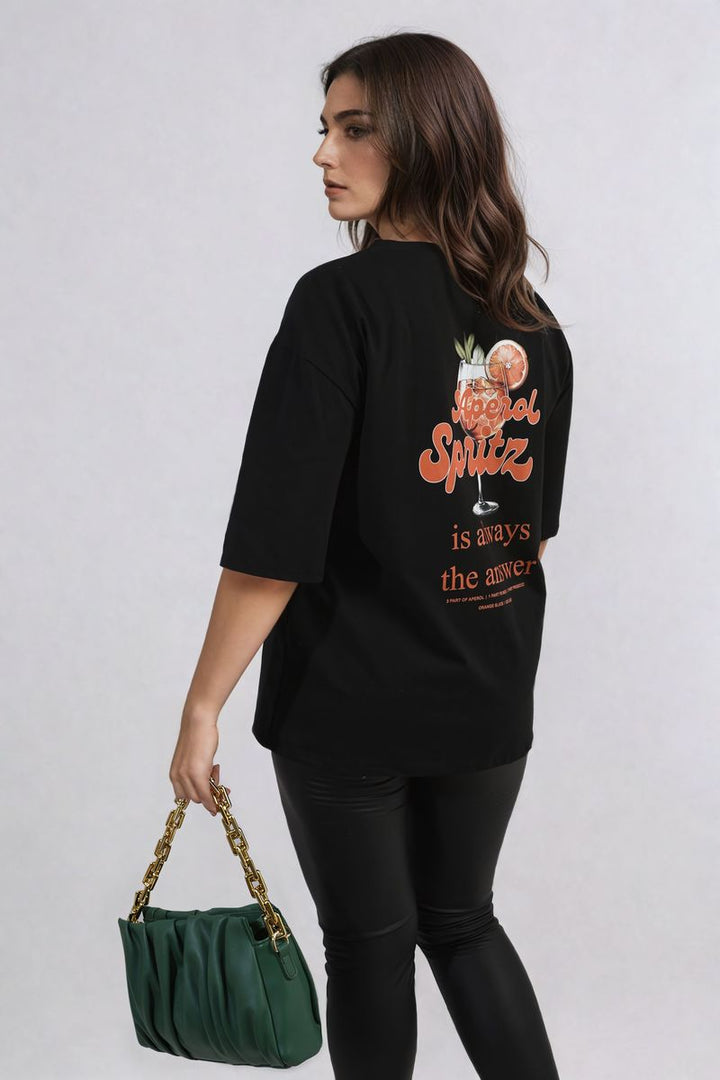 Woman wearing an Aperol Spritz Cocktail Graphic Top while holding a green purse with gold chains, embodying a casual, stylish look.