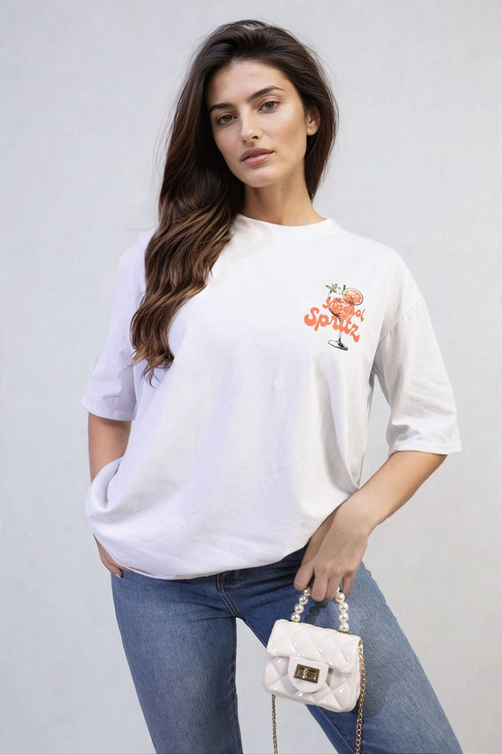 Woman in a white Aperol Spritz Cocktail Graphic Top, posing indoors, holding a white bag, embodying a casual and vibrant summer style.