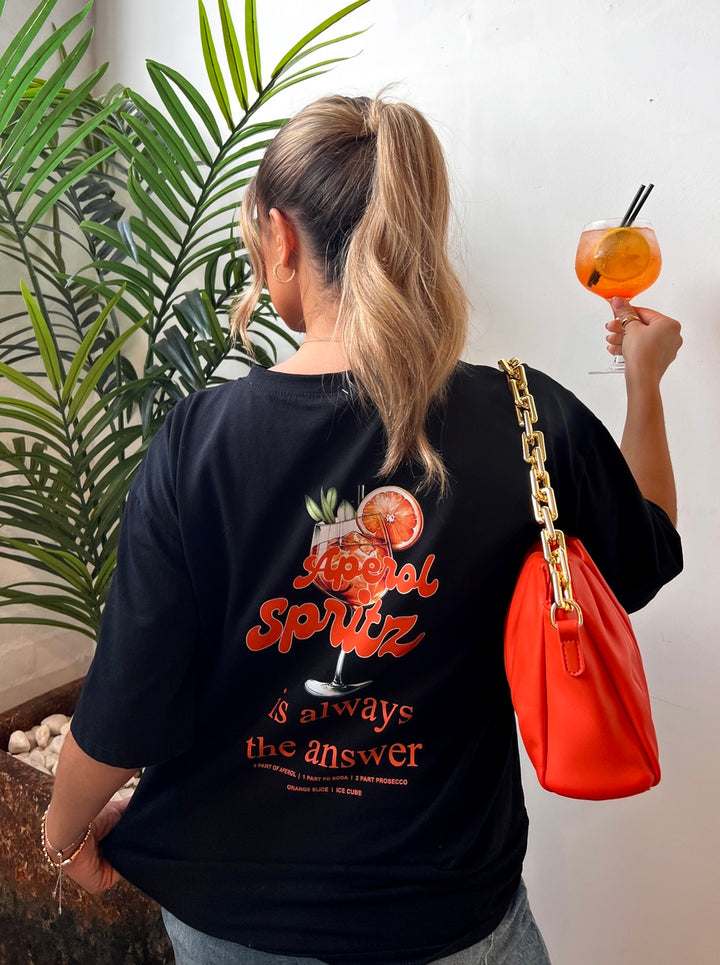 Woman holding drink, wearing Christina Aperol Spritz Cocktail Graphic Top, embodying vibrant summer vibes perfect for casual outings and beach days.