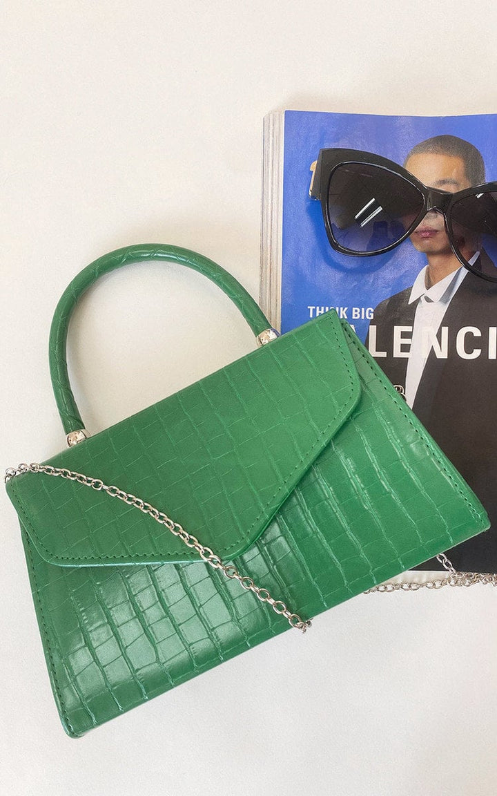 Croc Print Handbag with faux leather, featuring a handheld handle and chain strap, displayed alongside sunglasses on a magazine.