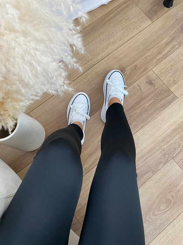 The photo captures an individual's legs clad in Moda's Stretchy Wide Waistband Second Skin Leggings in black, paired with white sneakers, standing on a light wood floor. Partially visible on the left is a fluffy decorative plant.