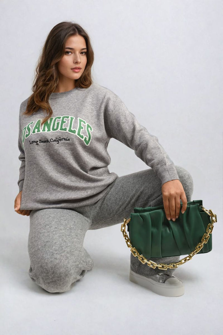 Woman wearing Los Angeles Graphic Long Sleeve Knitted Jumper, showcasing its trendy design and cozy fit. She holds a green purse, completing her stylish outfit.