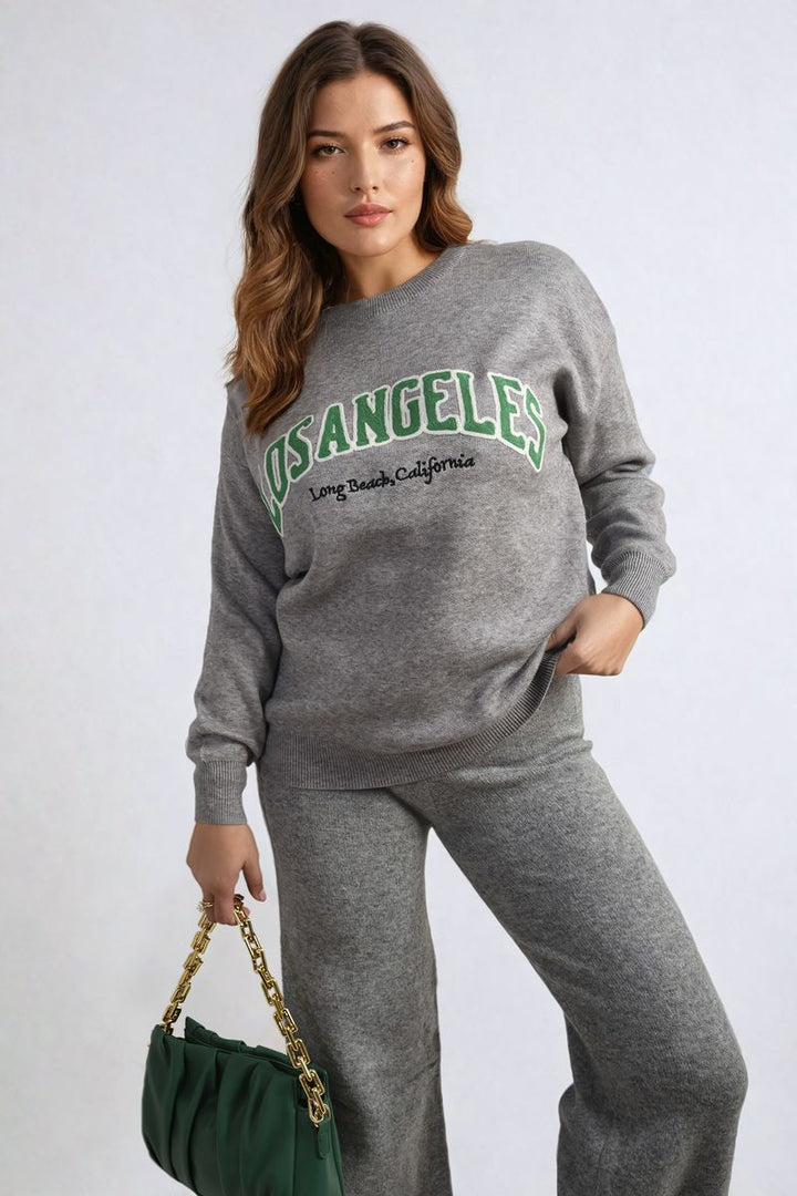 Woman wearing the Los Angeles Graphic Long Sleeve Knitted Jumper, featuring a trendy graphic, styled with grey pants and a green purse with a gold chain.