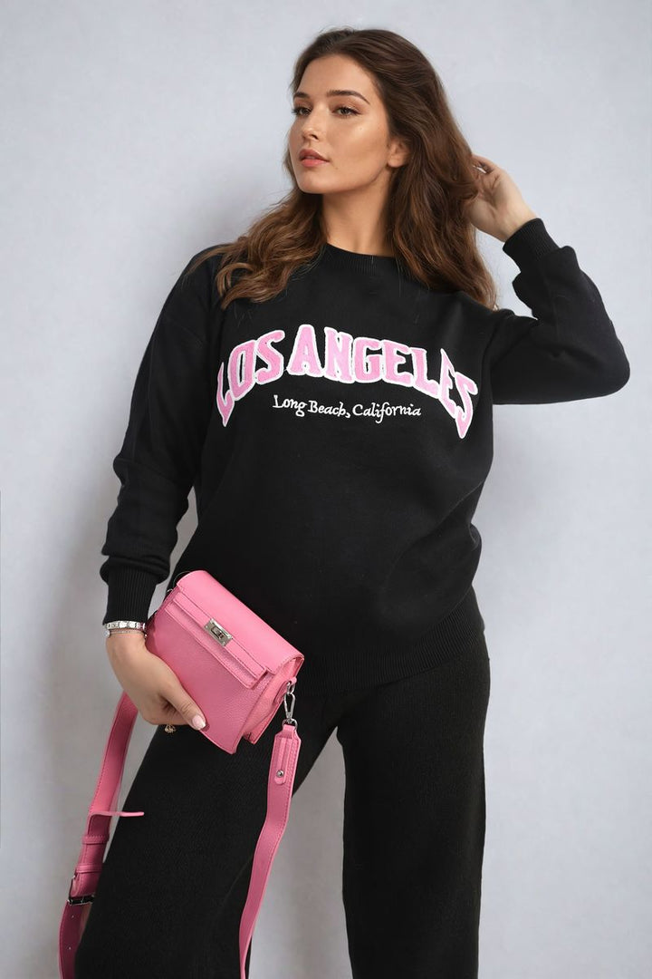 Woman wearing Los Angeles Graphic Long Sleeve Knitted Jumper, holding a pink purse. The jumper features a trendy Los Angeles graphic, perfect for stylish, everyday comfort.