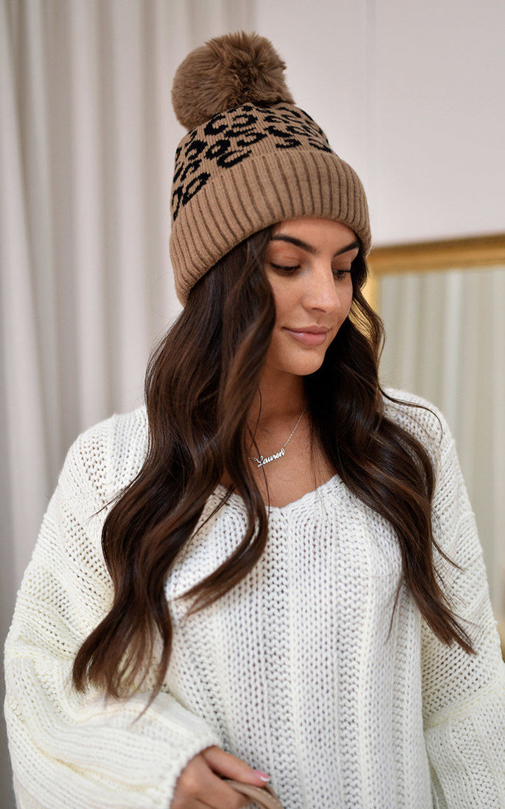 Woman wearing Edith Faux Fur Lined Beanie Leopard Hat with pom-pom detail, showcasing warm, stylish winter headwear.