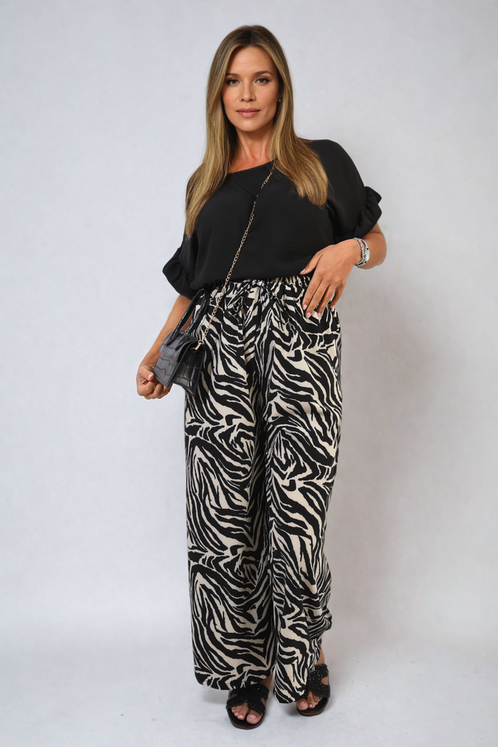 Woman wearing Elaina Zebra-Striped Print Wide Leg Trousers and black shirt, holding a black purse, highlighting stylish and versatile clothing from Holiday-clothes.co.uk.