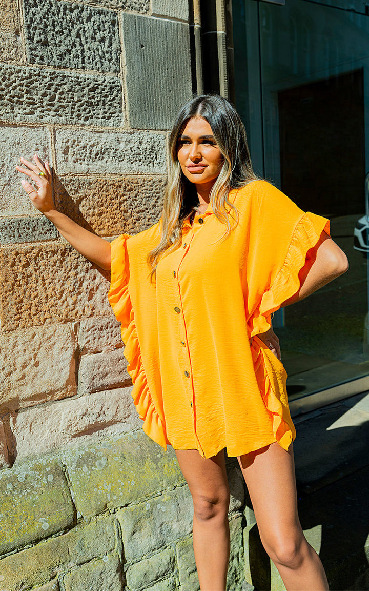 Woman in a Button Down Ruffle Sleeve Shirt Dress, leaning against a stone wall, showcasing the dress's ruffle detail and button-down front.