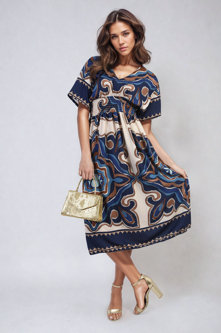 A woman in a Ruched Printed V-Neck Short Sleeve Midi Dress holds a gold purse, showcasing stylish holiday attire with gold sandals.