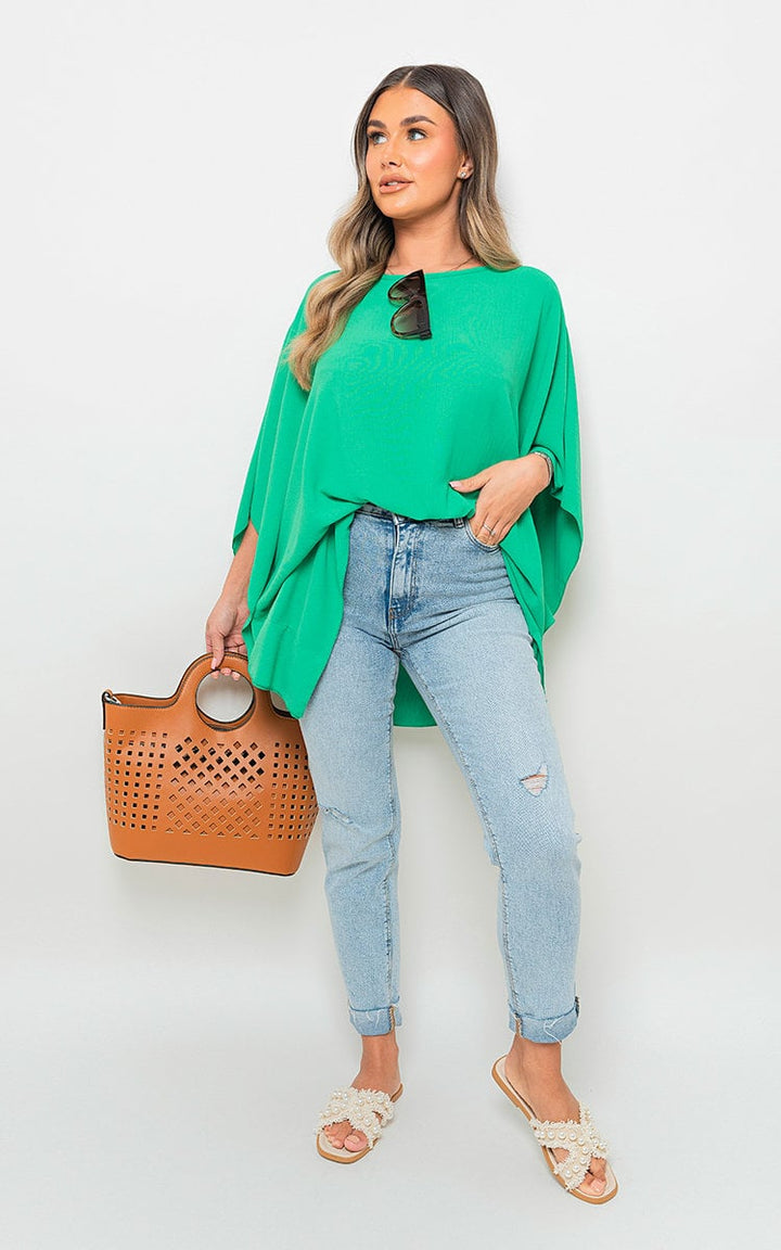 Woman in green oversized batwing sleeve casual top, holding a brown handbag, styled with blue jeans and white sandals.