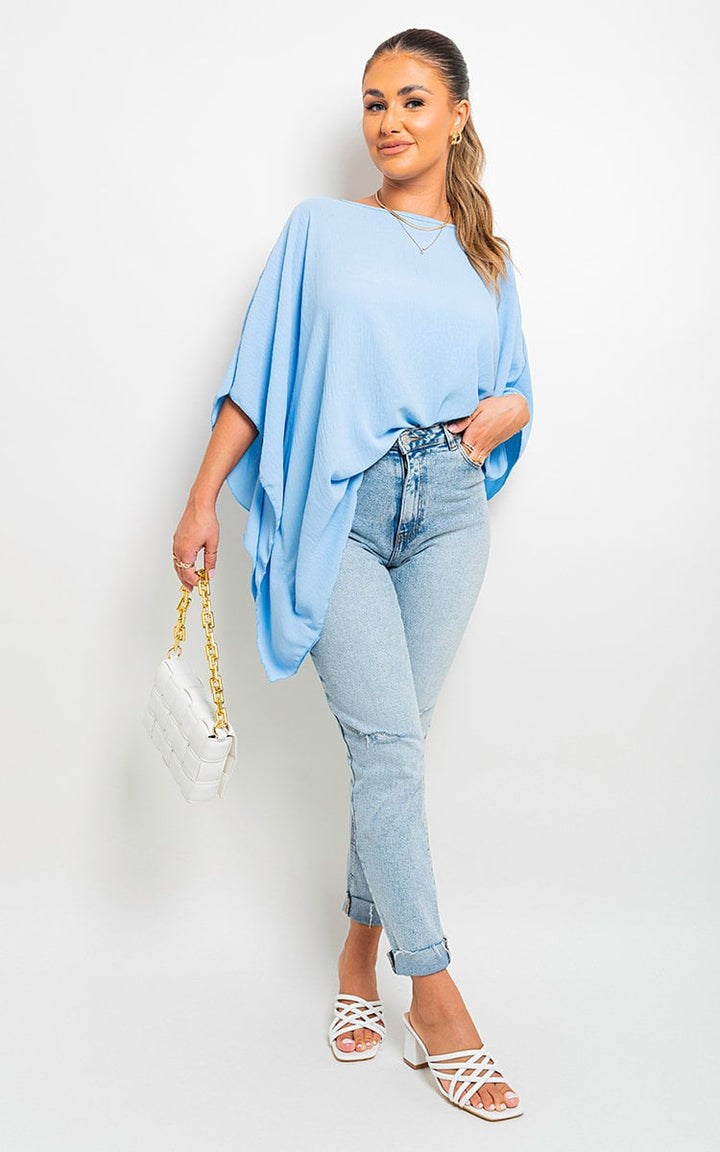 Woman in oversized batwing sleeve casual top, blue jeans, holding a white purse with gold chains, wearing white sandals. Fashionable, relaxed style from Holiday Clothes UK.