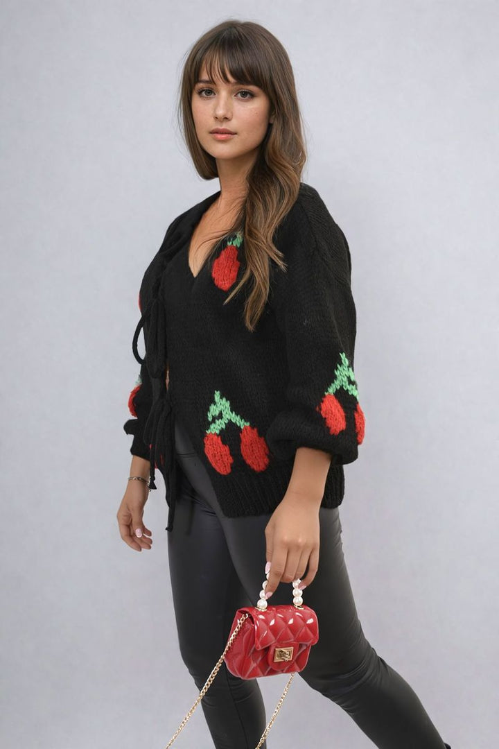 Woman wearing the Tie Front Cherry Print Knitted Cardigan, holding a red purse, showcasing the cardigan's playful cherry design and cozy knit style.