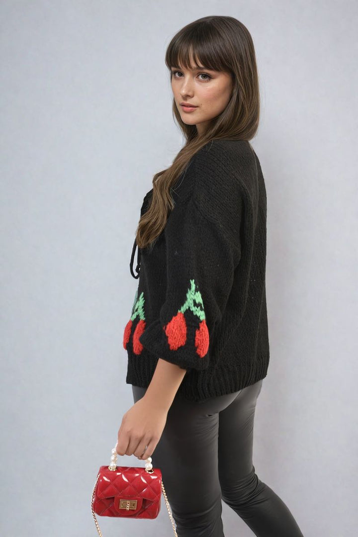 Woman in a black sweater holding a red purse, showcasing the Giselle Tie Front Cherry Print Knitted Cardigan, a cozy, playful addition to any wardrobe.