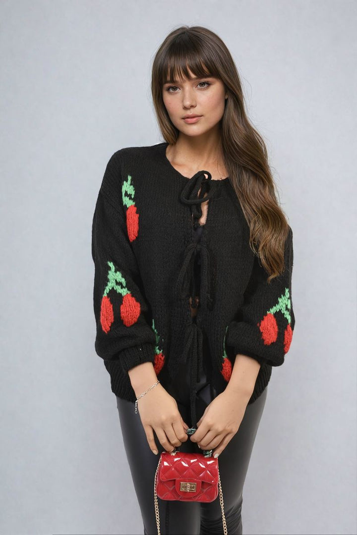 Woman wearing the Giselle Tie Front Cherry Print Knitted Cardigan, showcasing its playful cherry design and front tie detail, holding a red purse.