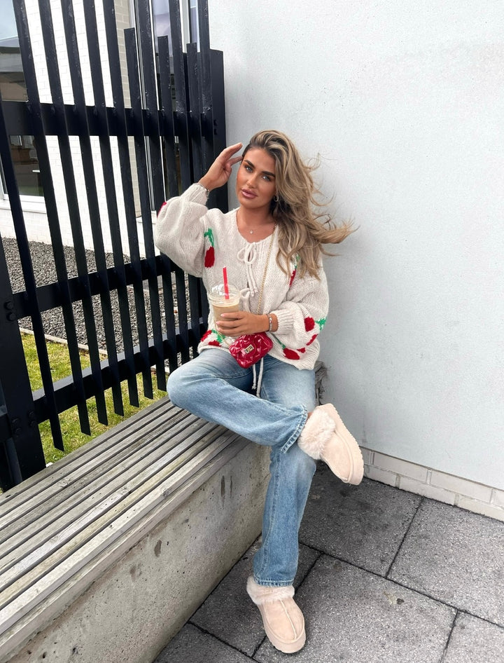 Woman sitting on a bench wearing the Giselle Tie Front Cherry Print Knitted Cardigan, showcasing its playful design and cozy knit.