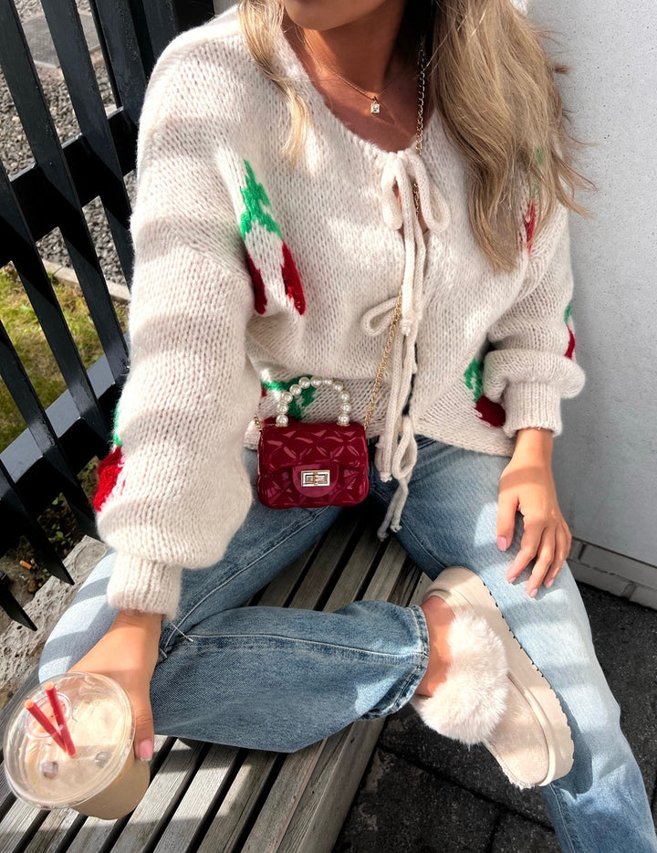 Woman wearing a Giselle Tie Front Cherry Print Knitted Cardigan, sitting on a bench. The cardigan features a charming cherry print and a playful tie front design.