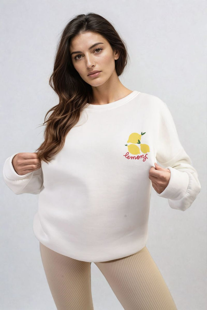 Woman wearing the Joyce Lemon Graphic Long Sleeve Knitted Jumper, featuring a cheerful lemon design, posing indoors. Cozy and stylish, perfect for a trendy look.