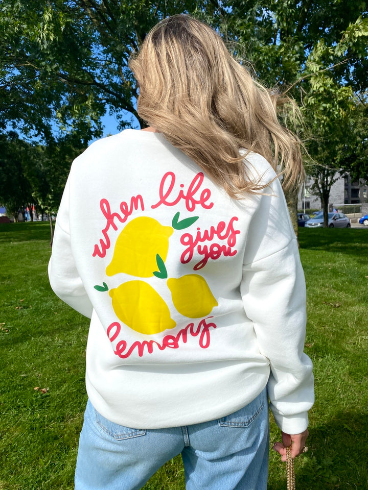 Person wearing the Joyce Lemon Graphic Long Sleeve Knitted Jumper, featuring a lemon design, paired with jeans in a park setting.