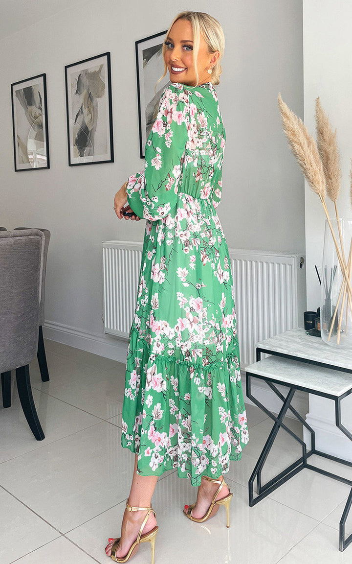 Woman wearing the Tiered Midi Dress with a green floral print, standing next to a marble table, showcasing a stylish and carefree summer look.