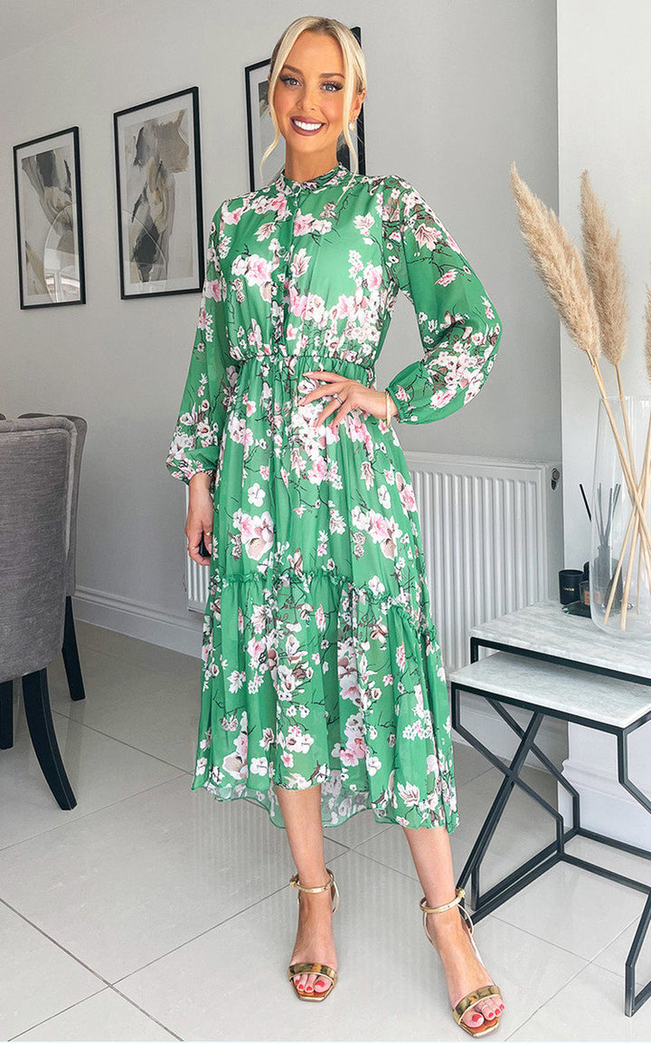 Woman in a green floral print Tiered Midi Dress, standing indoors, styled with gold sandals, reflecting a stylish and carefree summery look.