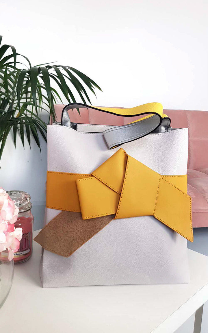 Colour Block Bow Tote Handbag with a prominent bow on front, featuring faux leather and a handheld strap, displayed against an indoor setting.