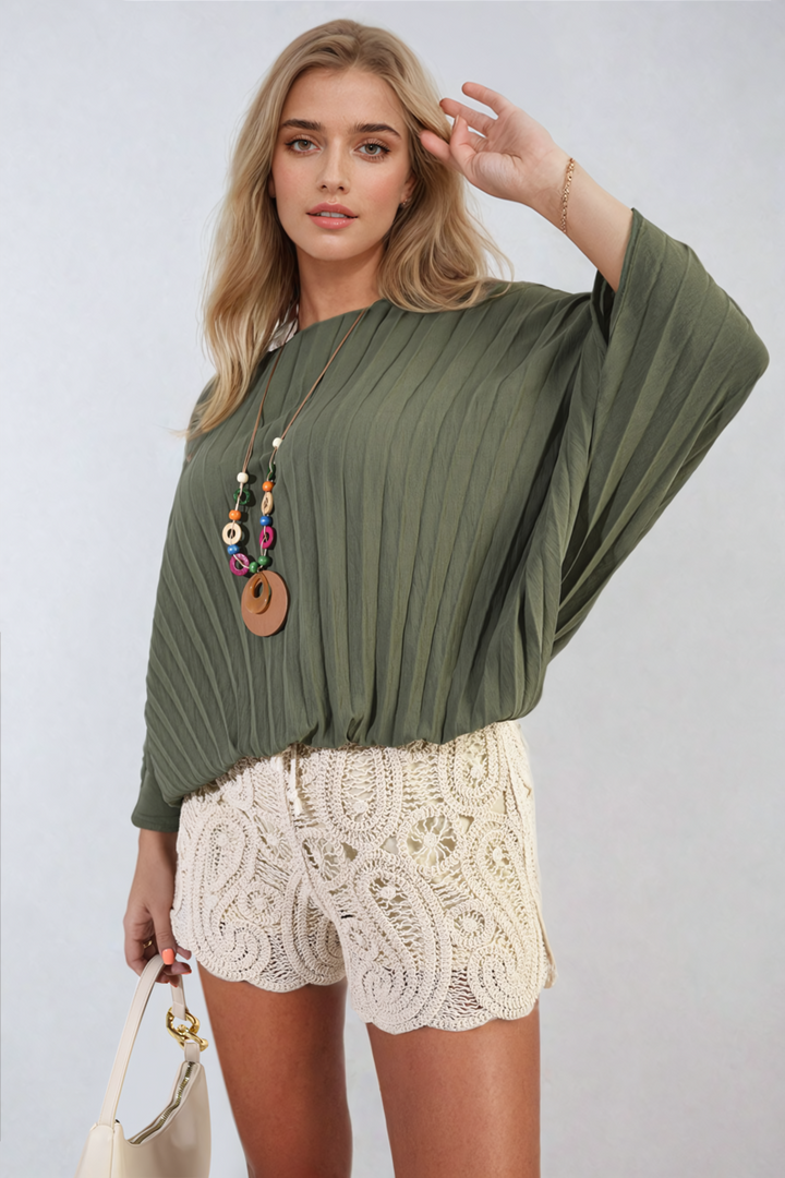 Woman wearing the Rowena Batwing Oversized Pleated Top, featuring dramatic batwing sleeves and modern pleats, paired with white shorts, embodying chic, versatile style.