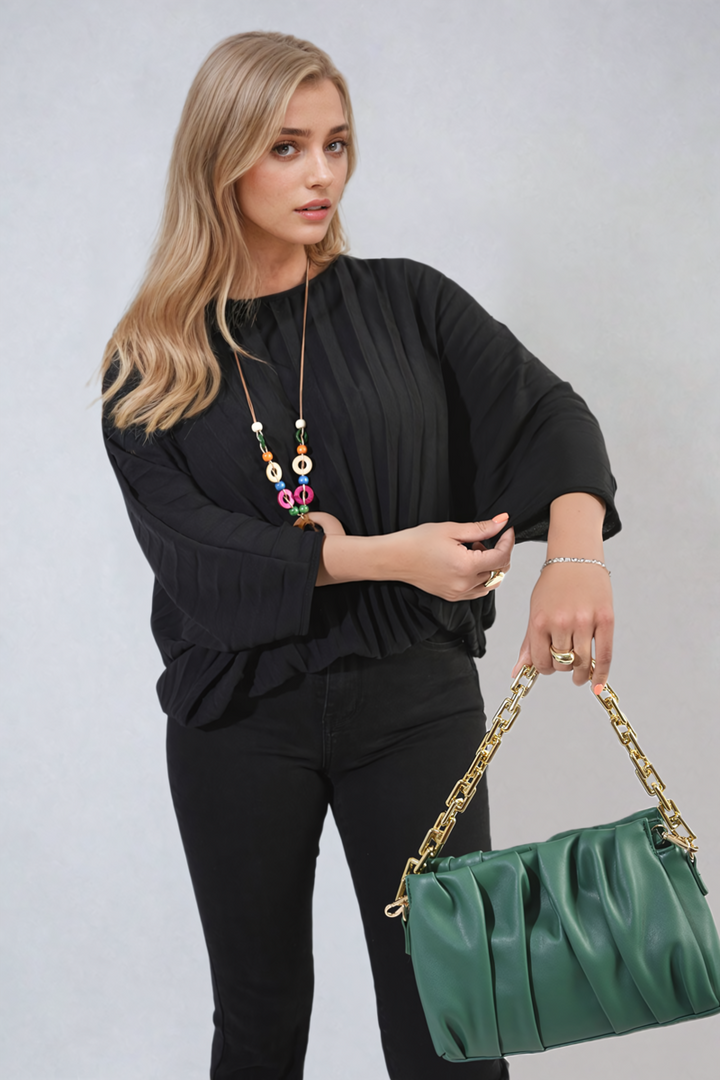 Woman showcasing a Batwing Oversized Pleated Top, paired with a green purse, highlighting the top's dramatic batwing sleeves and modern pleated design.