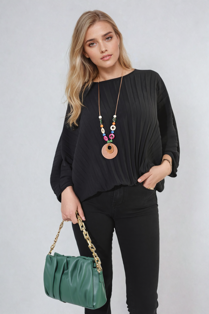 Woman in a black Rowena Batwing Oversized Pleated Top, accessorized with a green purse and necklace, showcasing a chic fashion ensemble.