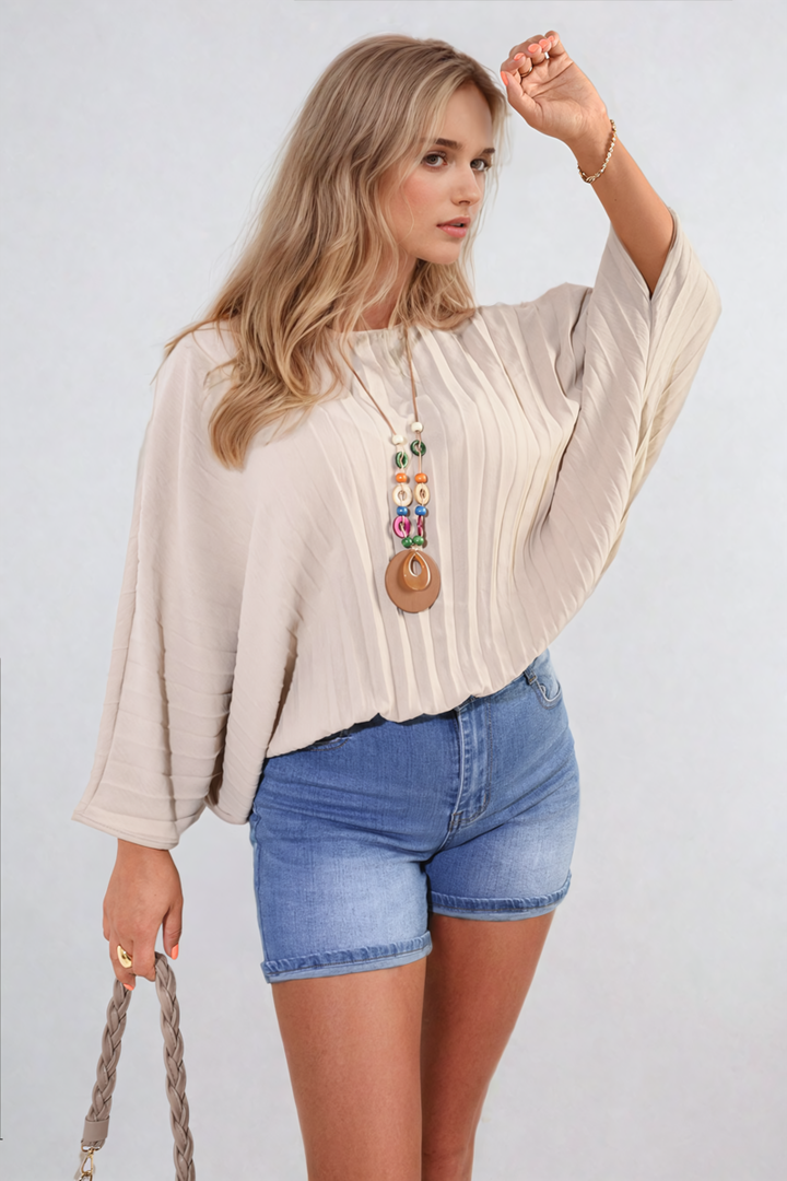 Woman wearing Rowena Batwing Oversized Pleated Top, showcasing batwing sleeves and pleated details, styled casually with blue shorts and a necklace.