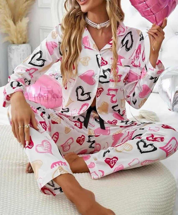 Woman in a heart print long sleeve button top and pants pyjamas set, sitting on a bed, showcasing cozy sleepwear from Holiday Clothes UK.