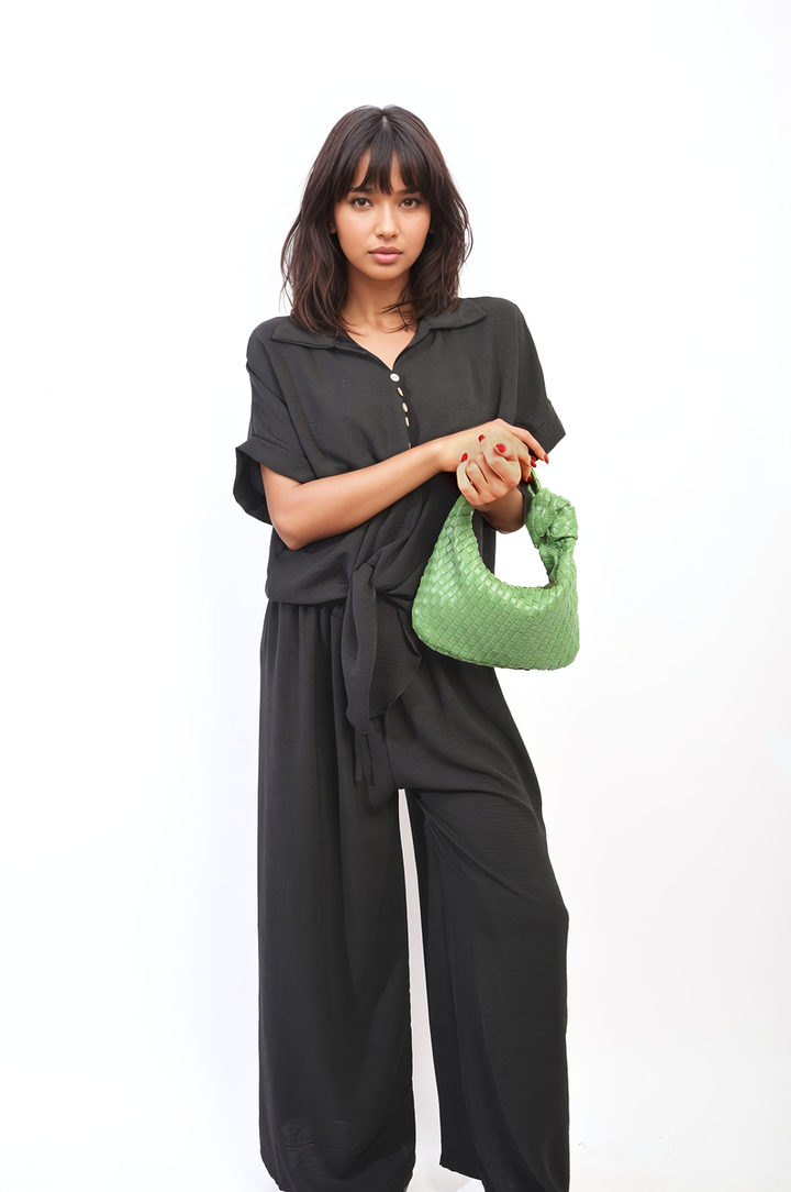 Woman in black co-ord set holding green purse, featuring a tie front top and wide leg pants.