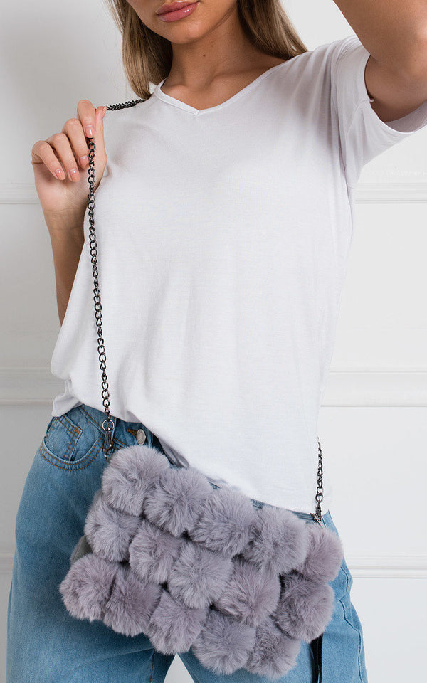 Woman in a white shirt and jeans holding the Cross Body Pom Pom Handbag, featuring pom pom detail, silver hardware, and a zip closure.