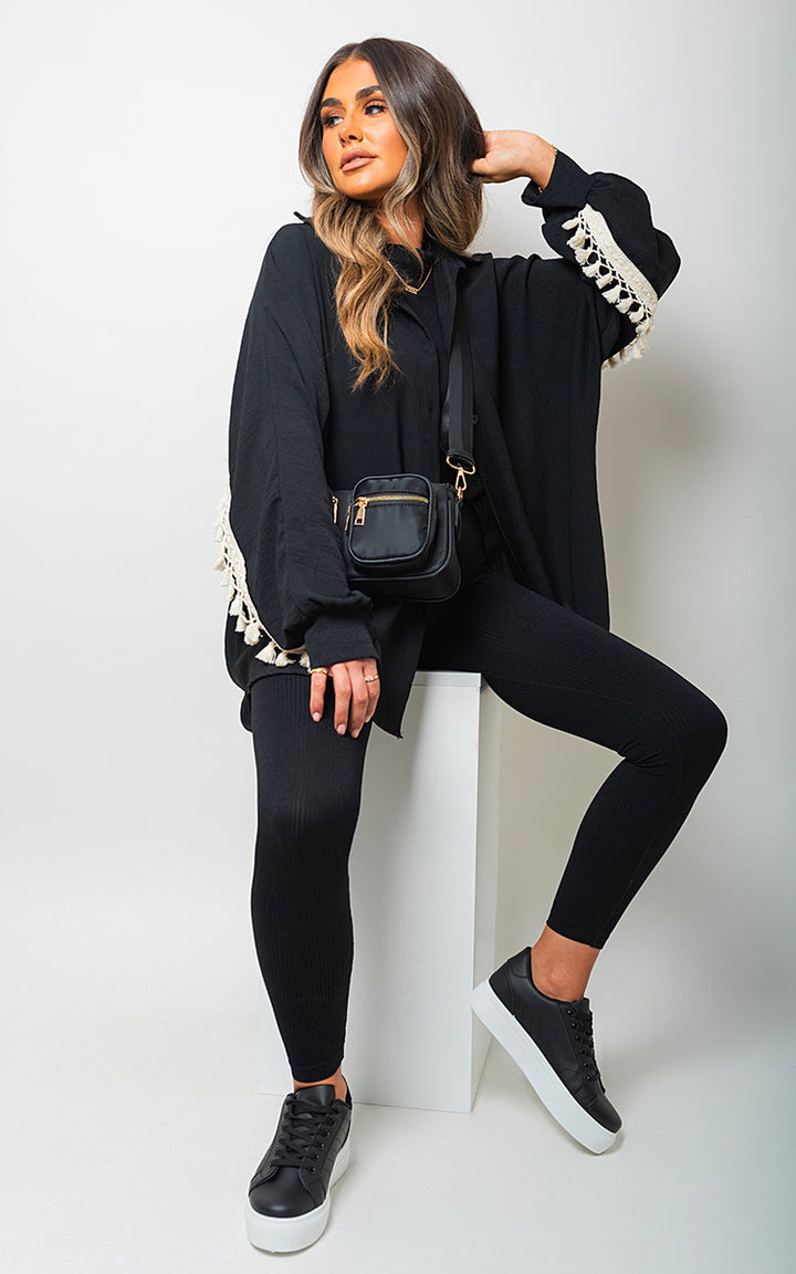 Woman wearing an Oversized Long Sleeve Tassel Trim Top, sitting on a white cube, showcasing boho chic style with a black shoulder bag and black and white shoes.