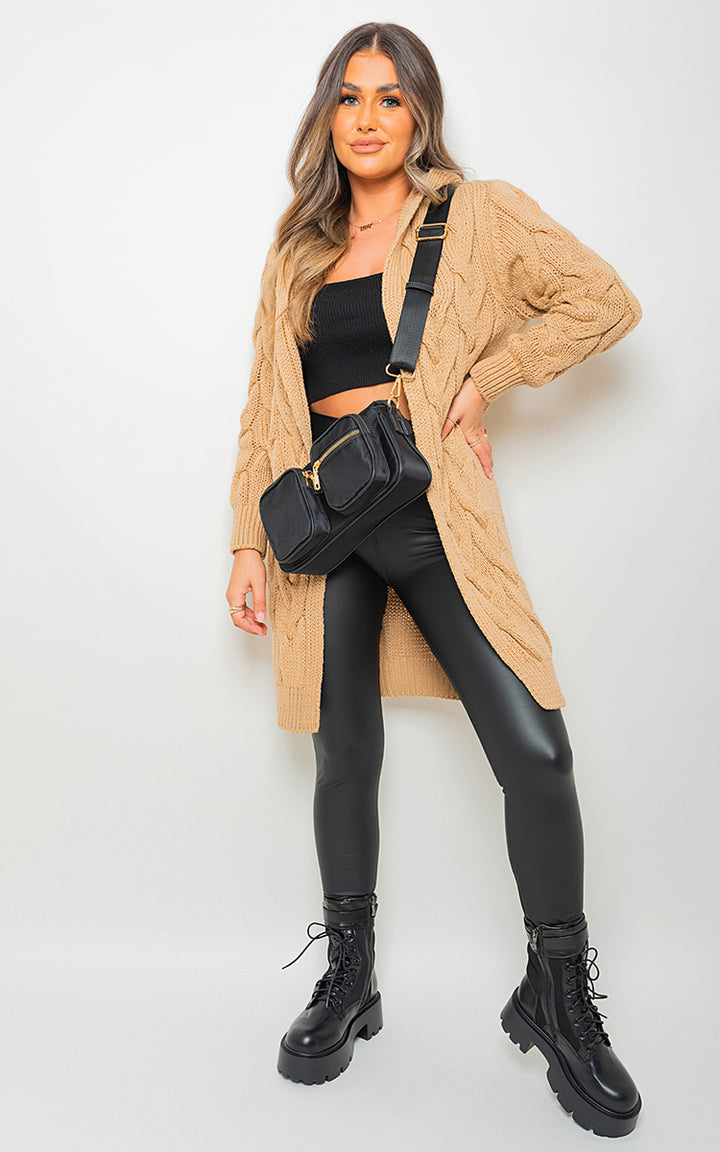 Woman wearing Chunky Cable Knitted Oversized Longline Hooded Cardigan, paired with black leather pants and boots, holding a black purse.