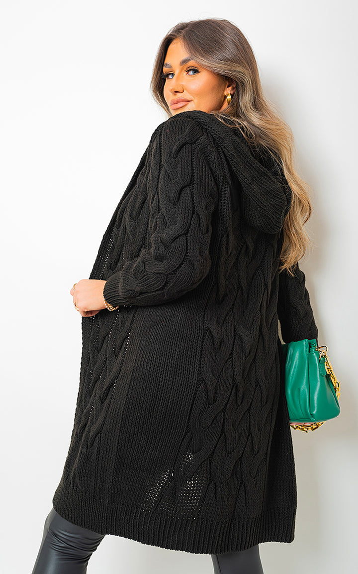 Woman in a black Chunky Cable Knitted Oversized Longline Hooded Cardigan, holding a green purse with a gold chain.