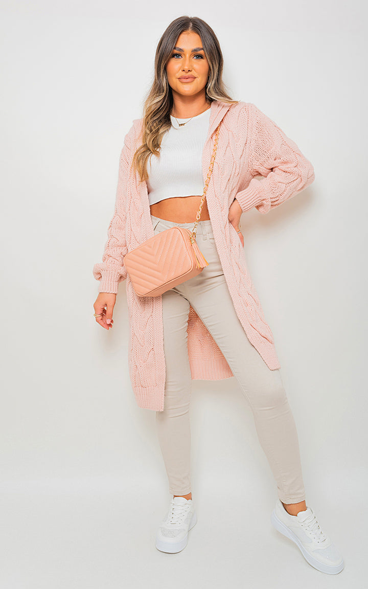 Woman wearing Chunky Cable Knitted Oversized Longline Hooded Cardigan, posing with a pink purse, styled for cozy layering.