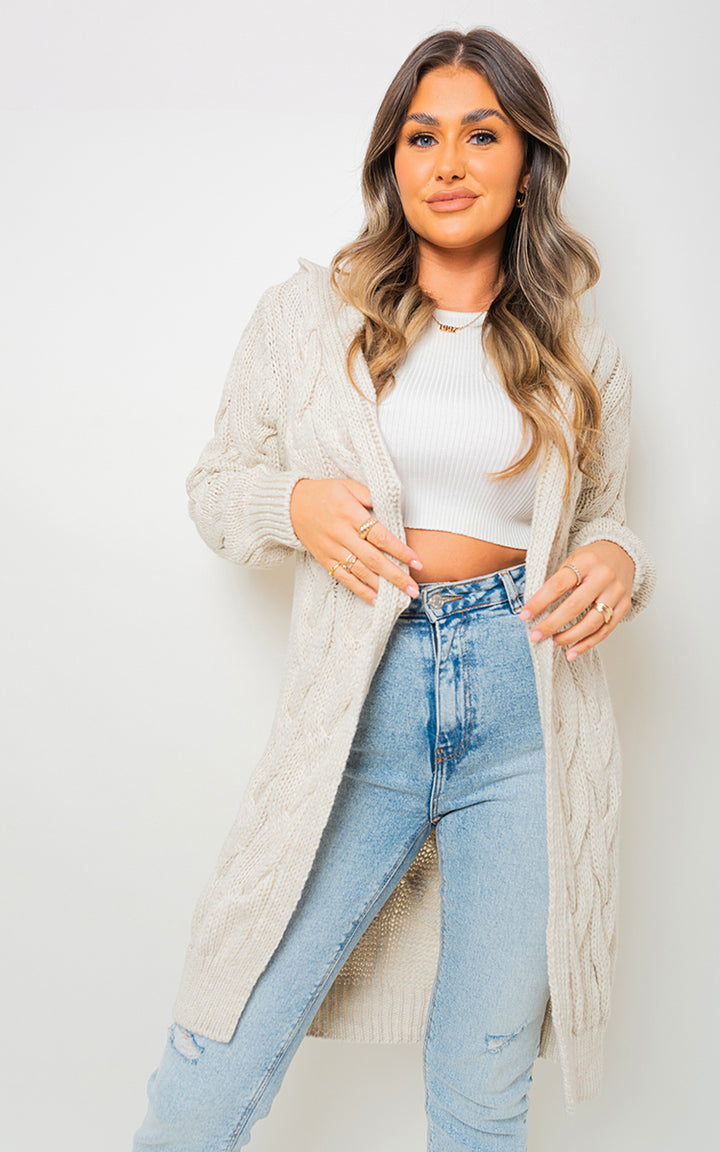 Woman wearing the Chunky Cable Knitted Oversized Longline Hooded Cardigan, posing indoors, highlighting its cozy, layered style. Suitable for casual, stylish occasions.