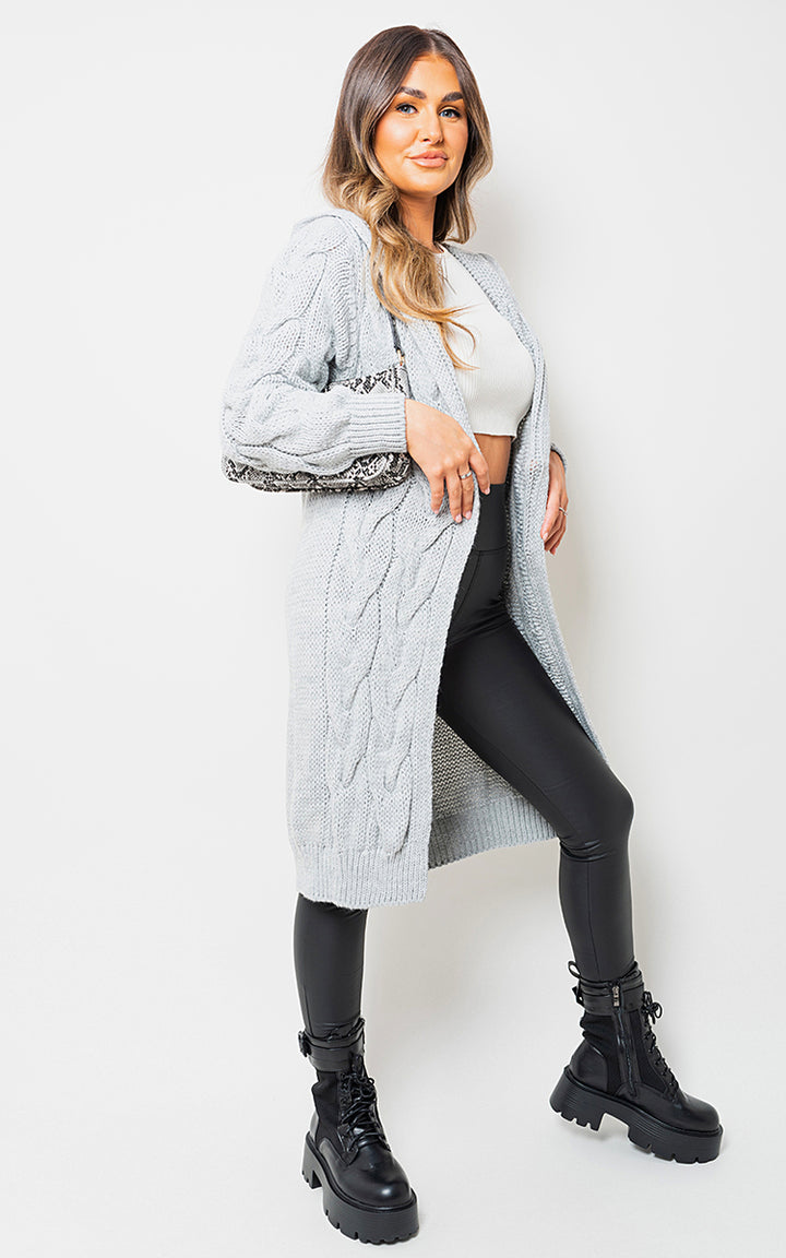 Woman wearing the Chunky Cable Knitted Oversized Longline Hooded Cardigan, posing, with a focus on fashion and comfort, alongside black boots for a stylish look.